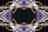 An intricate, symmetrical abstract pattern resembling a kaleidoscope, featuring swirling colors and shapes that appear fluid and dynamic. The design is centralized with elaborate loops and delicate lace-like details, blending hues of blue, purple, white, and gold.