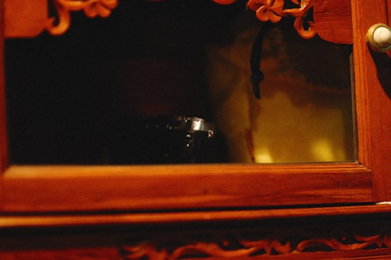 An old wooden cabinet partially displays a small, dark device, possibly a camera. The cabinet features intricate carvings and a brass knob. Warm light creates an ambient atmosphere.
