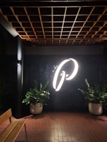 A large illuminated letter, possibly the letter 'P', is prominently displayed on a dark tiled wall. The scene includes two large potted plants on either side of the letter, positioned against a background of glossy black tiles under a wooden ceiling grid. A wooden bench is partially visible on the left.