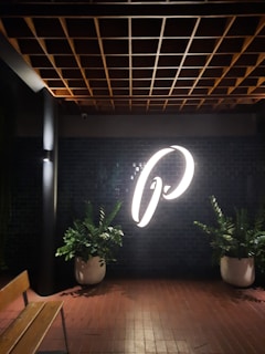 A large illuminated letter, possibly the letter 'P', is prominently displayed on a dark tiled wall. The scene includes two large potted plants on either side of the letter, positioned against a background of glossy black tiles under a wooden ceiling grid. A wooden bench is partially visible on the left.