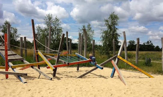 A playground area features a wooden climbing structure with colorful beams arranged diagonally, supported by a sandy ground. A child is climbing on the structure. In the background, there are wooden poles, green grass, and trees under a partly cloudy sky.