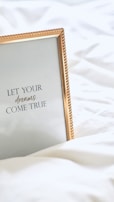 A framed inspirational quote on a white bedspread with a gold-colored frame. The quote reads 'Let your dreams come true' in elegant typography.