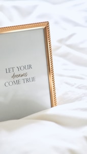 A framed inspirational quote on a white bedspread with a gold-colored frame. The quote reads 'Let your dreams come true' in elegant typography.