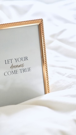 A framed inspirational quote on a white bedspread with a gold-colored frame. The quote reads 'Let your dreams come true' in elegant typography.