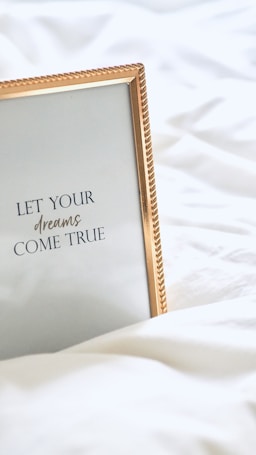 A framed inspirational quote on a white bedspread with a gold-colored frame. The quote reads 'Let your dreams come true' in elegant typography.