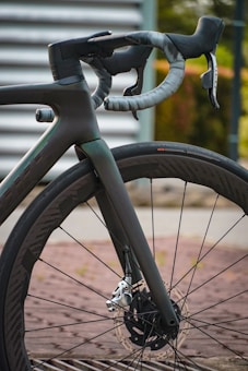 A close-up view of a sleek, modern road bike's front section, featuring the handlebar and front wheel. The handlebar is wrapped in gray tape with a streamlined design, and the front wheel has a disc brake rotor. The background includes a blurred metal and natural setting.