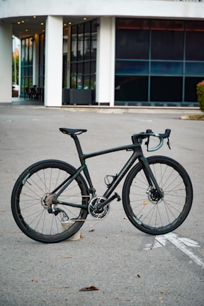 A sleek black bicycle stands on a paved surface in front of a modern building with large windows and white pillars. The bicycle has a lightweight frame, thin tires, and advanced gear mechanics.