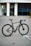 A sleek black bicycle stands on a paved surface in front of a modern building with large windows and white pillars. The bicycle has a lightweight frame, thin tires, and advanced gear mechanics.