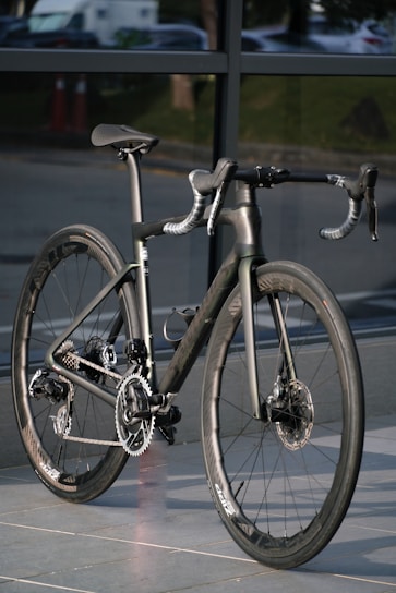 A sleek, modern road bike with aero handlebars and a carbon fiber frame, positioned against a background of glass windows. The sunlight highlights the bike's smooth surfaces and intricate gear system.