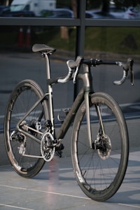 A sleek, modern road bike with aero handlebars and a carbon fiber frame, positioned against a background of glass windows. The sunlight highlights the bike's smooth surfaces and intricate gear system.