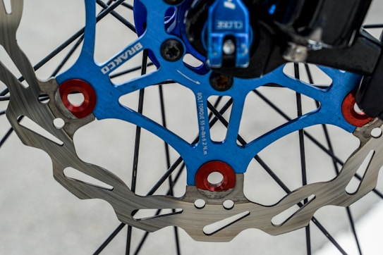 A close-up view of a bicycle brake disc. The disc features a metallic appearance with multiple spokes radiating from the center. The central area shows a blue and red design with text indicating bolt torque specifications. The disc is attached to a wheel with visible spokes, creating a mechanical and detailed aesthetic.