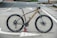 A high-end mountain bike is resting on its tires with the support of a can beneath the pedal. The bike is positioned on an empty road with marked lines visible on the asphalt. It features thick, knobby tires suitable for off-road terrain, a sleek black and gold frame, and a detailed gear system.