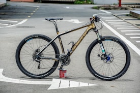 A high-end mountain bike is resting on its tires with the support of a can beneath the pedal. The bike is positioned on an empty road with marked lines visible on the asphalt. It features thick, knobby tires suitable for off-road terrain, a sleek black and gold frame, and a detailed gear system.