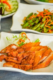 Slices of crispy, golden-brown fried meat are arranged on a white plate with a garnish of shredded vegetables. In the background, there are additional dishes, including a serving of stir-fried vegetables with thinly sliced carrots and whole green beans.