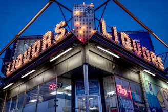 A corner liquor store with prominent neon signs, illuminated against a clear evening sky. The signs display the word 'LIQUORS' in bright, vintage-style bulbs. There are glass windows reflecting light and various neon signs, including one that reads 'OPEN'.