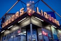 A corner liquor store with prominent neon signs, illuminated against a clear evening sky. The signs display the word 'LIQUORS' in bright, vintage-style bulbs. There are glass windows reflecting light and various neon signs, including one that reads 'OPEN'.