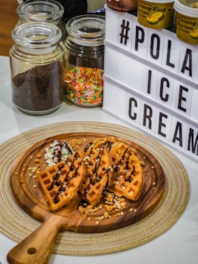 A wooden serving board holds freshly made waffles drizzled with chocolate sauce and a dollop of whipped cream. The board is placed on a woven placemat, flanked by glass jars containing chocolate sprinkles and colorful candy sprinkles. A lightbox sign displaying 'POLA ICE CREAM' is visible next to the jars, along with a few ice cream tubs.