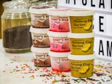 Various ice cream tubs are stacked in two columns. One column features Goat's Milk ice cream with flavors like chocolate and strawberry, while the other includes Musang King Durian ice cream. Accompanying the tubs are a jar of chocolate sprinkles and colorful sprinkles scattered on a white surface. A light-up board with partial text appears in the background.