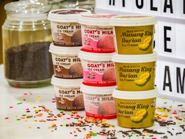 Various ice cream tubs are stacked in two columns. One column features Goat's Milk ice cream with flavors like chocolate and strawberry, while the other includes Musang King Durian ice cream. Accompanying the tubs are a jar of chocolate sprinkles and colorful sprinkles scattered on a white surface. A light-up board with partial text appears in the background.