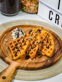 Three pieces of golden brown waffles are drizzled with chocolate syrup on a wooden serving board, accompanied by a swirl of whipped cream topped with chocolate sauce. Colorful sprinkles are visible in a jar in the background, along with part of a lightbox display.