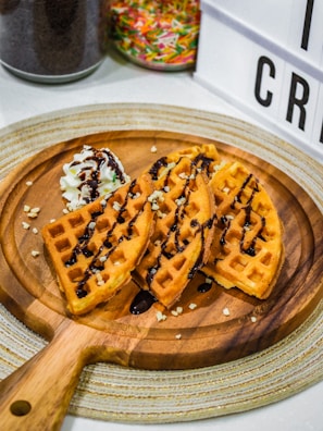 Three pieces of golden brown waffles are drizzled with chocolate syrup on a wooden serving board, accompanied by a swirl of whipped cream topped with chocolate sauce. Colorful sprinkles are visible in a jar in the background, along with part of a lightbox display.