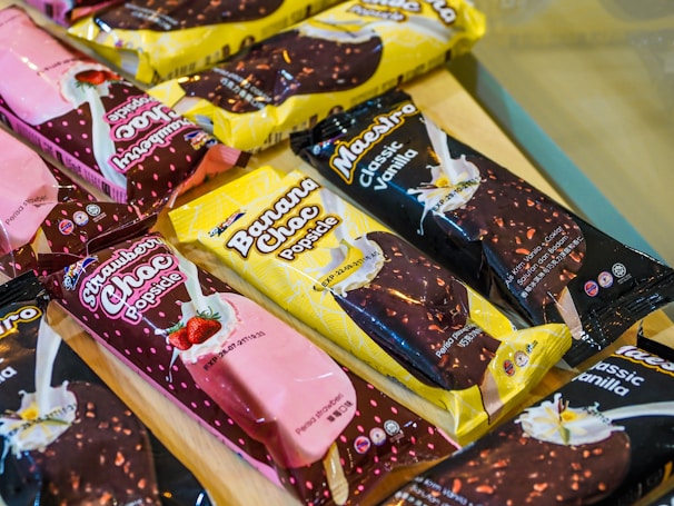 A collection of packaged ice cream popsicles displayed on a surface. The flavors visible include Strawberry Choc, Banana Choc, and Classic Vanilla. The packaging is colorful with branding and images of the respective flavors. The popsicles are arranged in an overlapping manner, combining shades of pink, yellow, and black.