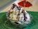A glass dish filled with scoops of colorful ice cream topped with whipped cream and drizzled with chocolate sauce. A small red umbrella is positioned as a decoration, and the dish rests on a textured green surface.