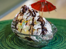 A glass dish filled with scoops of colorful ice cream topped with whipped cream and drizzled with chocolate sauce. A small red umbrella is positioned as a decoration, and the dish rests on a textured green surface.