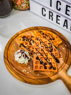 A wooden platter holds sliced waffles covered in chocolate sauce and sprinkled with small bits of nuts. There is a dollop of whipped cream beside the waffles. In the background, containers filled with colorful sprinkles are partially visible, next to a sign displaying the words 'ICE CREAM'.