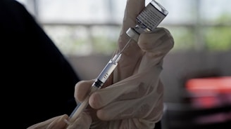 A gloved hand holds a syringe drawing liquid from a vial, likely involving a medical or vaccination procedure. The background is blurred, putting emphasis on the careful and precise action being undertaken.