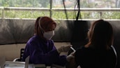 A healthcare worker wearing a mask and gloves administers a vaccine to a person sitting across from them. There is a backdrop of greenery, partially visible through a window or barrier, creating a soft and muted atmosphere.