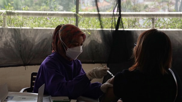 A healthcare worker wearing a mask and gloves administers a vaccine to a person sitting across from them. There is a backdrop of greenery, partially visible through a window or barrier, creating a soft and muted atmosphere.