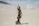 A bronze statue of Lady Justice holding a balance scale in her right hand and a sword in her left hand. She wears a blindfold and stands with a book under her foot. The background is blurred, with a light blue sky and a flat beige surface.
