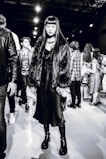 A stylish individual wearing a long dark dress, chunky platform boots, and a faux fur coat stands confidently. The background reveals several people milling around, suggesting a busy event or fashion show atmosphere.