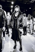 A stylish individual wearing a long dark dress, chunky platform boots, and a faux fur coat stands confidently. The background reveals several people milling around, suggesting a busy event or fashion show atmosphere.