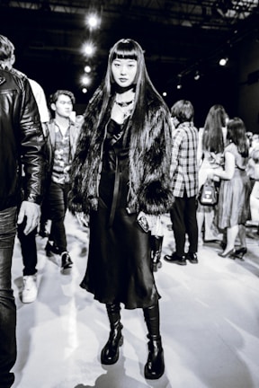 A stylish individual wearing a long dark dress, chunky platform boots, and a faux fur coat stands confidently. The background reveals several people milling around, suggesting a busy event or fashion show atmosphere.