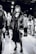 A stylish individual wearing a long dark dress, chunky platform boots, and a faux fur coat stands confidently. The background reveals several people milling around, suggesting a busy event or fashion show atmosphere.