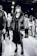 A stylish individual wearing a long dark dress, chunky platform boots, and a faux fur coat stands confidently. The background reveals several people milling around, suggesting a busy event or fashion show atmosphere.