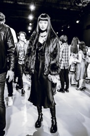 A stylish individual wearing a long dark dress, chunky platform boots, and a faux fur coat stands confidently. The background reveals several people milling around, suggesting a busy event or fashion show atmosphere.