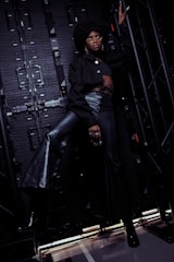 A person with an afro hairstyle and glasses is posing confidently against an industrial backdrop, wearing a black crop top and high-waisted, lace-up leather pants. The scene is dimly lit, emphasizing dramatic shadows and textures.