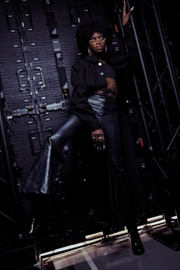 A person with an afro hairstyle and glasses is posing confidently against an industrial backdrop, wearing a black crop top and high-waisted, lace-up leather pants. The scene is dimly lit, emphasizing dramatic shadows and textures.