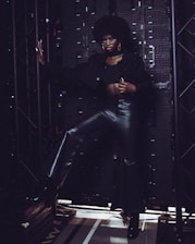 A person stands confidently in front of an industrial backdrop, dressed in a stylish outfit consisting of a black top, leather pants, and high-heeled boots. The individual's pose is dynamic, with one hand resting on a metal framework and the other on their hip. The background is composed of dark metal panels and structures, creating a contrasting setting.