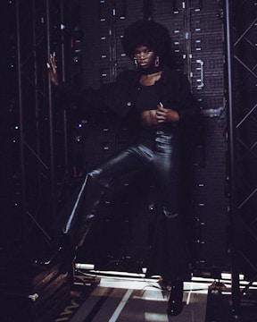 A person stands confidently in front of an industrial backdrop, dressed in a stylish outfit consisting of a black top, leather pants, and high-heeled boots. The individual's pose is dynamic, with one hand resting on a metal framework and the other on their hip. The background is composed of dark metal panels and structures, creating a contrasting setting.