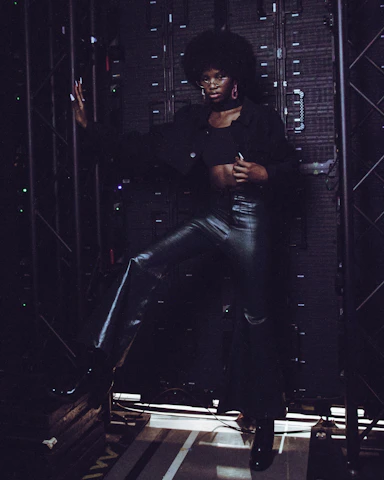 A striking, fashion-forward female domme in a sleek, elegant outfit, posed confidently against a moody, film-inspired backdrop.