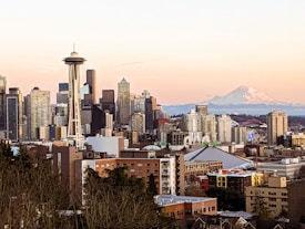An urban skyline with numerous high-rise buildings and the iconic Space Needle prominently featured. In the background, a snow-capped mountain, possibly Mount Rainier, adds a dramatic backdrop. The scene is captured during a sunset, with warm tones in the sky.