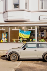 A gray car is parked on the street in front of a building with large windows. A Ukrainian flag is displayed in one of the windows, alongside decorative yellow flowers. The building appears to be a business, with signs in German.