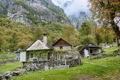 Nestled in a picturesque valley, a rustic mountain village features small stone cottages surrounded by lush greenery and towering cliffs. Mist shrouds parts of the rocky landscape, adding a mystical quality. The cottages, with their quaint architecture and wooden elements, are partially covered in vibrant foliage, suggesting an idyllic and serene rural life.