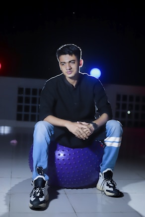 A person is seated on a purple exercise ball in a dimly lit area with red and blue lights in the background. They are wearing a black shirt, light blue jeans, and sneakers. The setting appears to be indoors, with a tiled floor and panels suggesting windows or a grid pattern.