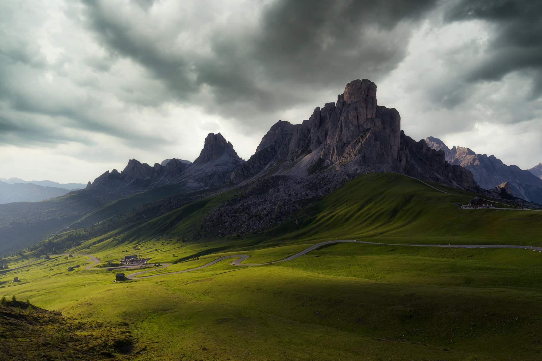Photograph of Dark storm clouds curl above the green road bends and rocky summit of Passo Giau.