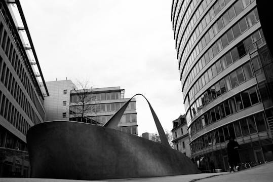 A modern urban landscape featuring a large, abstract metal sculpture positioned between two contemporary high-rise buildings. The sculpture curves upward with sleek lines, contrasting against the glass and steel architecture surrounding it. A person is walking nearby, adding a sense of scale to the scene. The overcast sky creates a stark black and white visual.